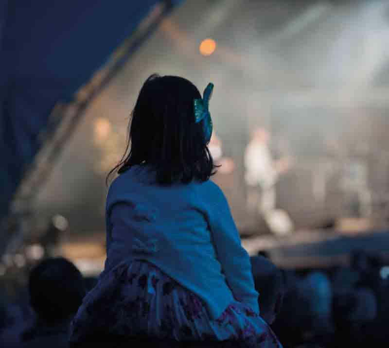 A young girl sits on her Fathers shoulders watching the acts on stage.