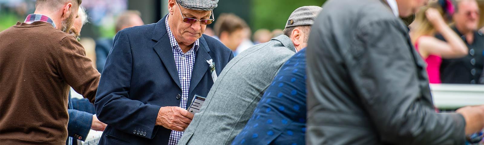 Group of gentlemen at the races, with one reading through a racecard.