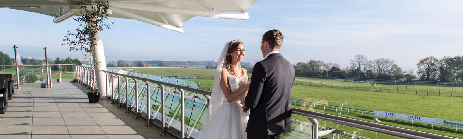 A bride and groom smile lovingly at each other in the roof garden at Bath racecourse.